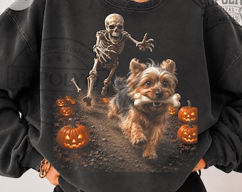 Puede incluir: Una sudadera de cuello redondo gris carbón con un estampado gráfico de un perro pequeño marrón y negro corriendo con un hueso en la boca, perseguido por un esqueleto humano. La escena incluye varias calabazas naranjas brillantes.