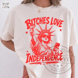 May include: A cream-colored short-sleeve t-shirt featuring a red graphic of the Statue of Liberty wearing sunglasses, holding a martini glass and a smartphone. The text BITCHES LOVE INDEPENDENCE is printed in red above and below the illustration.