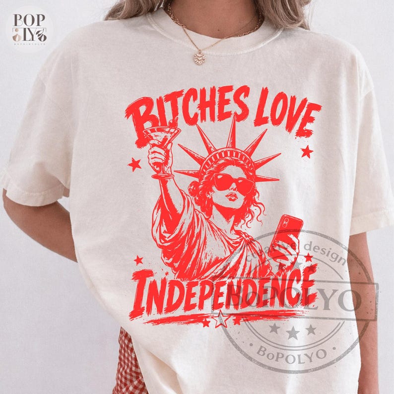 May include: A cream-colored short-sleeve t-shirt featuring a red graphic of the Statue of Liberty wearing sunglasses, holding a martini glass and a smartphone. The text BITCHES LOVE INDEPENDENCE is printed in red above and below the illustration.