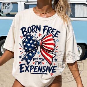 May include: Cream-colored t-shirt with the text "BORN FREE" above a bow design in red, white, and blue, with the text "BUT NOW I'M EXPENSIVE". The design includes stars and hearts.