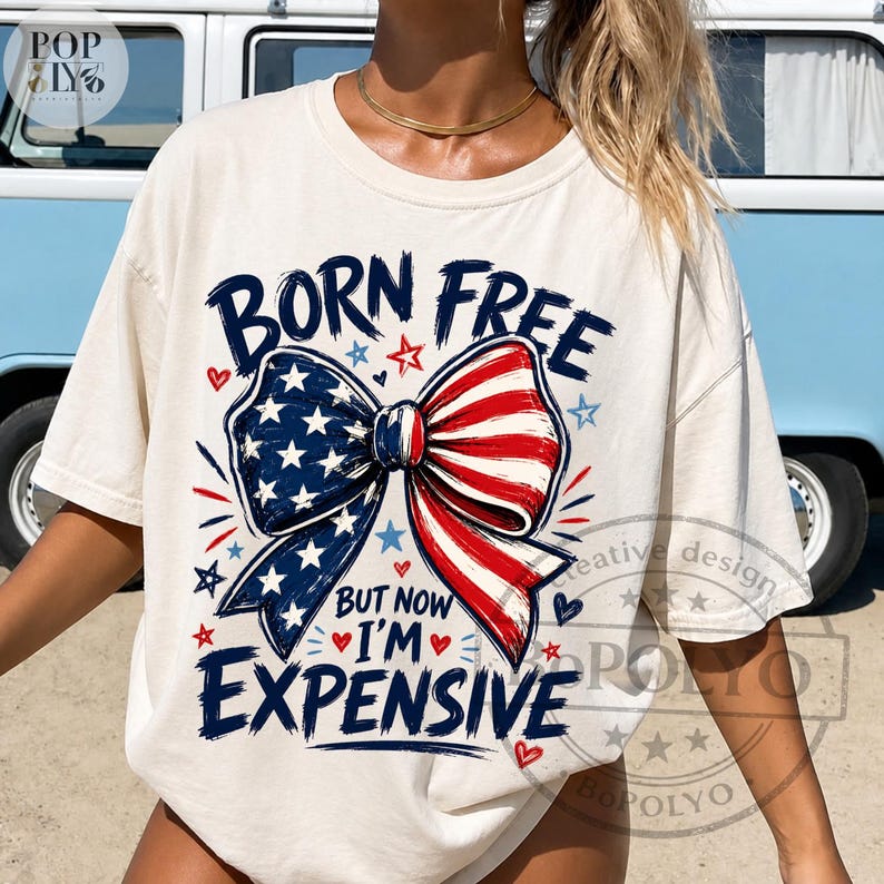 May include: Cream-colored t-shirt with the text "BORN FREE" above a bow design in red, white, and blue, with the text "BUT NOW I'M EXPENSIVE". The design includes stars and hearts.