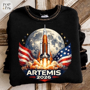 Op de afbeelding: Zwarte sweatshirt met een kleurrijke afbeelding van een raket die naar de maan lanceert, geflankeerd door Amerikaanse vlaggen. Het ontwerp bevat de tekst "ARTEMIS 2026" in het wit. De sweatshirt heeft een geribbelde kraag.