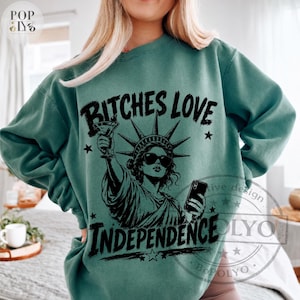 May include: A teal crewneck sweatshirt featuring a black graphic print of the Statue of Liberty wearing sunglasses, holding a martini glass in one hand and a smartphone in the other, with the text BITCHES LOVE INDEPENDENCE printed in black.
