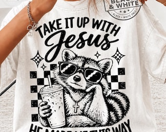 May include: A cream-colored t-shirt featuring a black graphic of a raccoon wearing sunglasses and a cross necklace, holding a drink. The text reads "TAKE IT UP WITH JESUS" and "HE MADE ME THIS WAY" with a checkered background pattern.