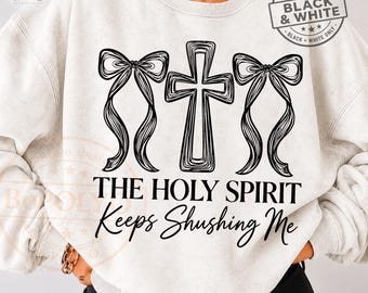 May include: A light gray crewneck sweatshirt featuring a black graphic of a central cross flanked by two bows. Below the graphic, the text reads THE HOLY SPIRIT Keeps Shushing Me. A circular badge in the top right corner says TWO COLOR BLACK & WHITE.