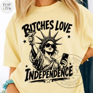May include: A pale yellow t-shirt featuring a black graphic print of the Statue of Liberty wearing sunglasses, holding a martini glass in one hand and a smartphone in the other, with the text BITCHES LOVE INDEPENDENCE printed above and below.