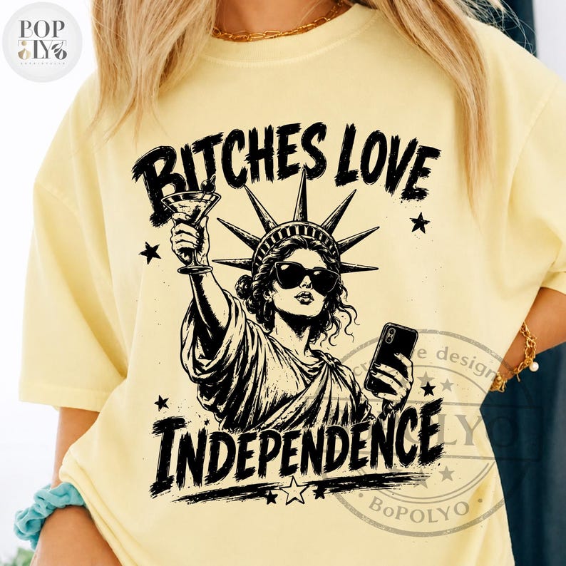 May include: A pale yellow t-shirt featuring a black graphic print of the Statue of Liberty wearing sunglasses, holding a martini glass in one hand and a smartphone in the other, with the text BITCHES LOVE INDEPENDENCE printed above and below.
