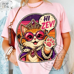 Könnte beinhalten: Rosa T-Shirt mit einer Cartoon-Katze, die eine Sonnenbrille, eine Krone und einen Umhang trägt. Die Katze winkt und eine Sprechblase sagt "Hi Zev!" Das Design hat einen Retro-Sonnenuntergangshintergrund.