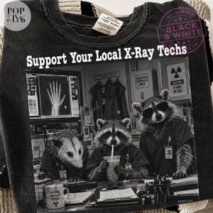 Może przedstawiać: Czarny t-shirt z grafiką przedstawiającą trzy szopy pracze w pracowni radiologicznej. Napis głosi Support Your Local X-Ray Techs. Jeden szop nosi okulary przeciwsłoneczne i strój medyczny, drugi pije z kubka, a trzeci siedzi przy monitorze z rentgenem dłoni. Na pierwszym planie znajduje się kubek z napisem Caffeine Contrast Repeat.