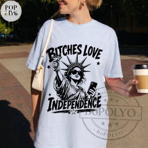 May include: A light blue t-shirt featuring a black graphic print of the Statue of Liberty wearing sunglasses, holding a martini glass and a smartphone. The text BITCHES LOVE INDEPENDENCE is printed in a bold, distressed black font above and below.