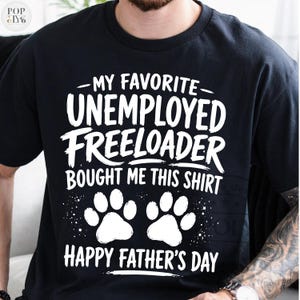 Könnte beinhalten: Schwarzes T-Shirt mit weißem Text: "MY FAVORITE UNEMPLOYED FREELOADER BOUGHT ME THIS SHIRT" und "HAPPY FATHER'S DAY". Mit zwei weißen Pfotenabdrücken. Ein lässiges, humorvolles Design.