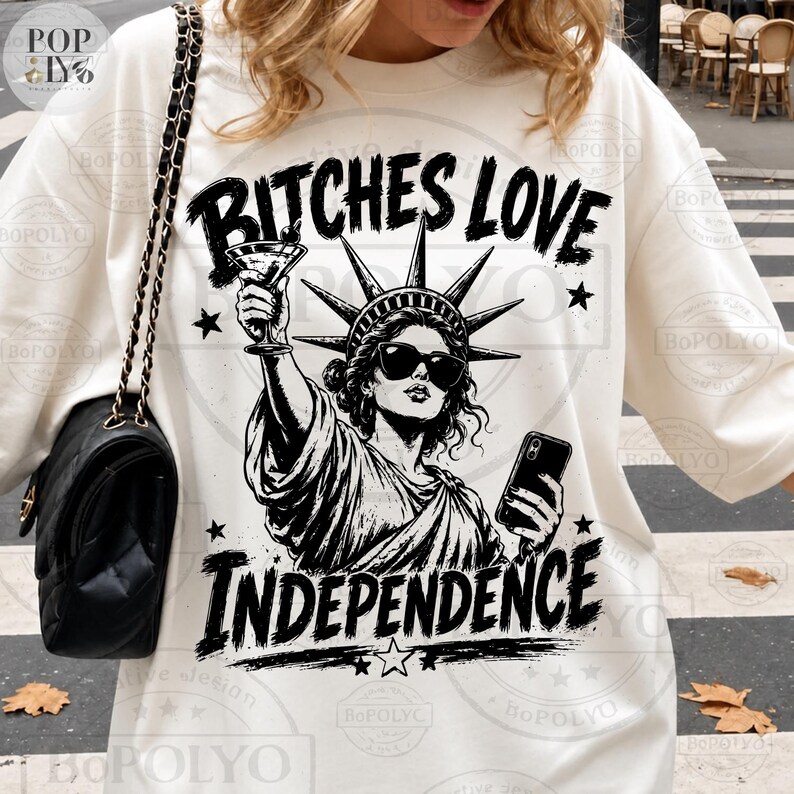 May include: An off-white t-shirt featuring a black graphic of the Statue of Liberty wearing sunglasses, holding a martini glass in one hand and a smartphone in the other, with the text BITCHES LOVE INDEPENDENCE printed in a bold, distressed font.