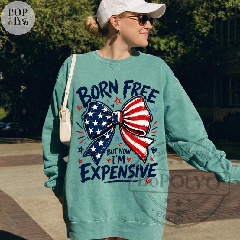 May include: Teal-colored sweatshirt with the text "BORN FREE BUT NOW I'M EXPENSIVE" and a patriotic bow design in red, white, and blue. The bow features stars and stripes, evoking an American theme.