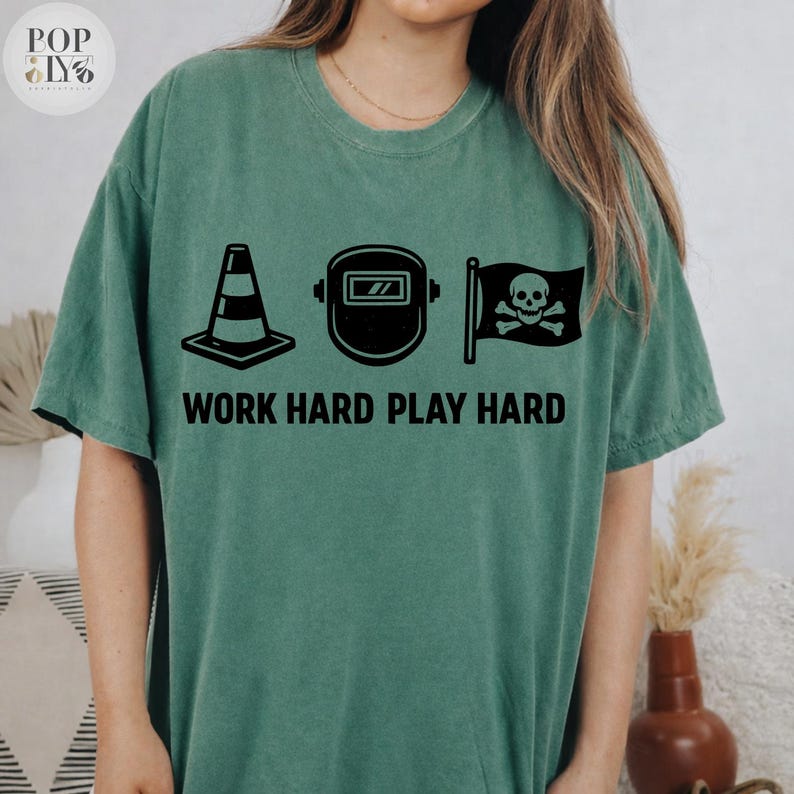 May include: Green t-shirt featuring black graphics: a traffic cone, a welding mask, and a skull and crossbones flag. The text "WORK HARD PLAY HARD" is printed below. The shirt has a loose fit.