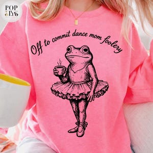 Puede incluir: Sudadera rosa con la frase "Off to commit dance mom foolery". La imagen muestra una ilustración en blanco y negro de una rana con tutú sosteniendo una taza de café.