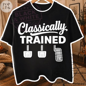 Pode incluir: Uma t-shirt preta de manga curta com o texto branco Classically TRAINED sobre uma ilustração gráfica de três pedais de carro, incluindo embraiagem, travão e acelerador, dispostos em fila na parte frontal da peça.