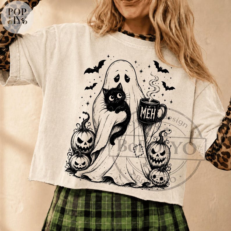 Puede incluir: Una camiseta de manga corta color crema con un estampado gr&aacute;fico negro de un fantasma sosteniendo un gato negro y una taza humeante con la palabra MEH. El dise&ntilde;o incluye tres calabazas en la base y varios murci&eacute;lagos volando.