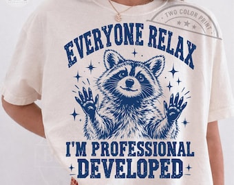 May include: A cream-colored short-sleeve t-shirt featuring a dark blue graphic of a raccoon with its paws raised. The text reads EVERYONE RELAX I'M PROFESSIONAL DEVELOPED. The design includes small star accents surrounding the raccoon illustration.