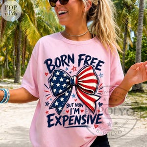 May include: A light pink t-shirt with the text "BORN FREE" above a bow tie design in red, white, and blue. The bow tie has a pattern of stars and stripes. The text "BUT NOW I'M EXPENSIVE" is below the bow tie.