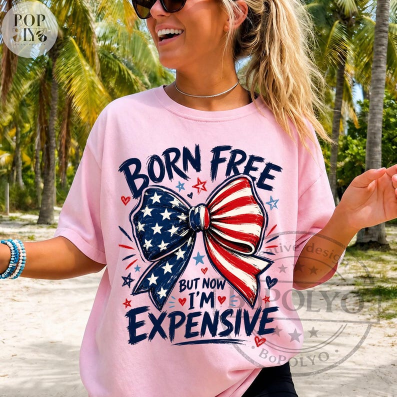 May include: A light pink t-shirt with the text "BORN FREE" above a bow tie design in red, white, and blue. The bow tie has a pattern of stars and stripes. The text "BUT NOW I'M EXPENSIVE" is below the bow tie.