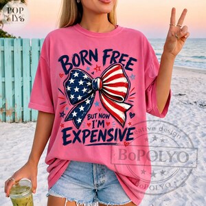 May include: Pink t-shirt with the text "BORN FREE BUT NOW I'M EXPENSIVE" and a bow design featuring the American flag. The shirt has short sleeves and a relaxed fit. The design includes stars, hearts, and a patriotic color scheme.