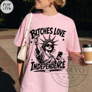 May include: A light pink short-sleeve t-shirt featuring a black graphic of the Statue of Liberty wearing sunglasses, holding a martini glass and a smartphone. The text BITCHES LOVE INDEPENDENCE is printed in a bold black font above and below the graphic.