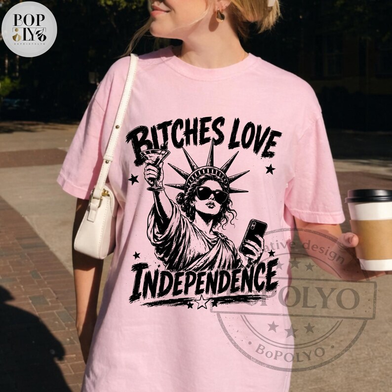May include: A light pink short-sleeve t-shirt featuring a black graphic of the Statue of Liberty wearing sunglasses, holding a martini glass and a smartphone. The text BITCHES LOVE INDEPENDENCE is printed in a bold black font above and below the graphic.