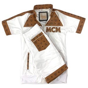 Puede incluir: Conjunto de chaqueta deportiva blanca de manga corta y pantalones cortos a juego con detalles marrones y el logo MCM. La chaqueta tiene cierre de cremallera y el texto "The Jacket Xpert". Los pantalones cortos tienen un bolsillo marrón.
