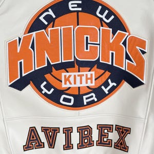 May include: A white leather jacket featuring a large circular New York Knicks basketball logo in orange and navy blue with the text KITH in the center. Below the logo, the word AVIREX is embroidered in orange and navy blue block lettering.