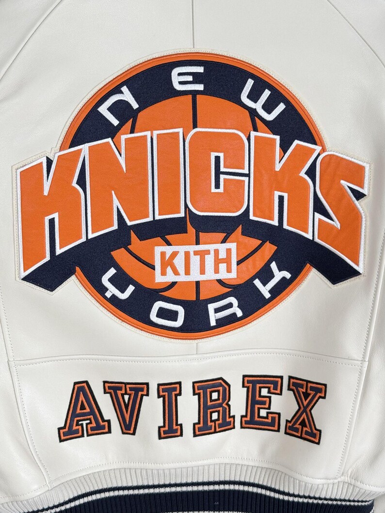 May include: A white leather jacket featuring a large circular New York Knicks basketball logo in orange and navy blue with the text KITH in the center. Below the logo, the word AVIREX is embroidered in orange and navy blue block lettering.