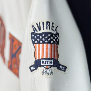 May include: A white leather jacket sleeve featuring an embroidered patch. The patch displays the text AVIREX at the top, a shield with stars and orange and white stripes, the text KITH in the center, N.Y. on the sides, and the year 1975 below.