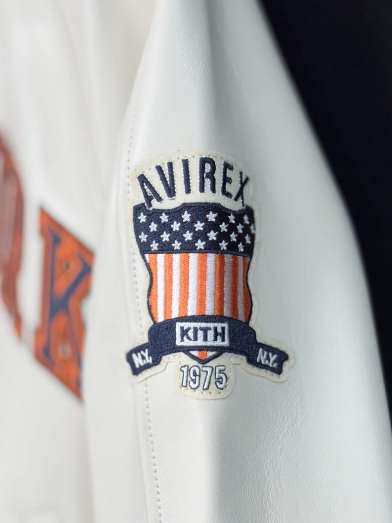 May include: A white leather jacket sleeve featuring an embroidered patch. The patch displays the text AVIREX at the top, a shield with stars and orange and white stripes, the text KITH in the center, N.Y. on the sides, and the year 1975 below.