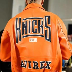 May include: A bright orange leather varsity jacket featuring the word KNICKS in large dark blue letters across the back, with the word KITH in a white box below it and the word AVIREX in dark blue letters near the bottom ribbed hem.