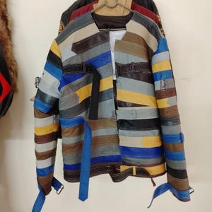 Op de afbeelding: Een veelkleurige leren jas met een patchwork-ontwerp. De jas heeft horizontale strepen in verschillende kleuren, waaronder blauw, geel, bruin en grijs. Het heeft een kraagloos ontwerp, meerdere riemen en een unieke, opvallende stijl.