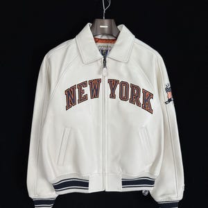May include: A white leather jacket featuring a full-length front zipper, a classic collar, and ribbed cuffs and hem with navy blue and white stripes. The chest displays the text NEW YORK in navy blue and orange lettering with an embroidered patch.