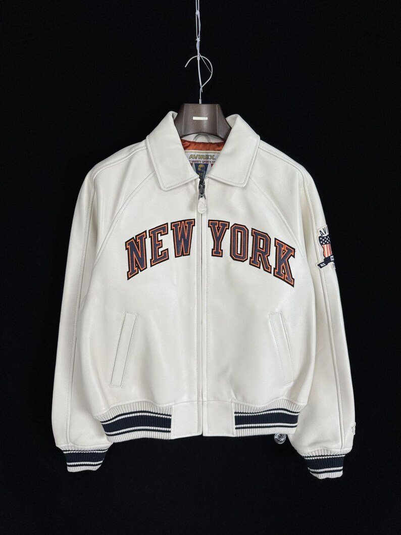 May include: A white leather jacket featuring a full-length front zipper, a classic collar, and ribbed cuffs and hem with navy blue and white stripes. The chest displays the text NEW YORK in navy blue and orange lettering with an embroidered patch.