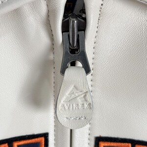 May include: A white leather jacket featuring a metal zipper pull with the embossed text AVIREX. A matching white leather pull tab is attached to the zipper, displaying an embossed AVIREX logo. Below the zipper, parts of orange and navy blue lettering are visible.