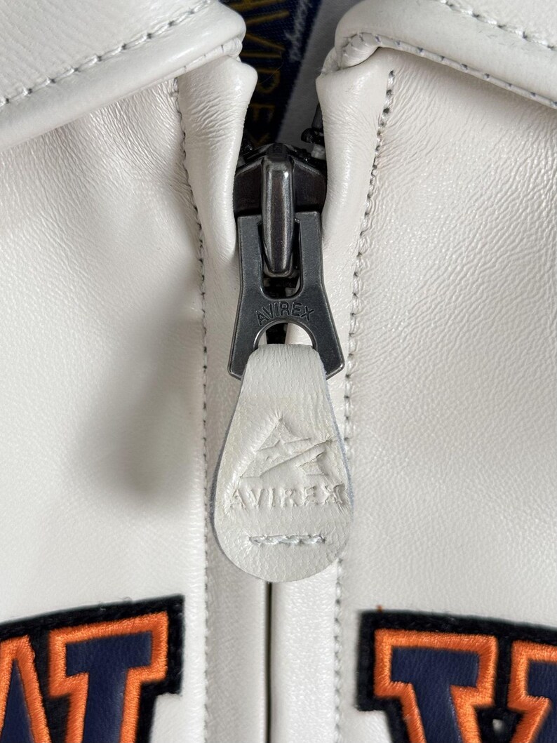 May include: A white leather jacket featuring a metal zipper pull with the embossed text AVIREX. A matching white leather pull tab is attached to the zipper, displaying an embossed AVIREX logo. Below the zipper, parts of orange and navy blue lettering are visible.