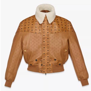 Op de afbeelding: Een bruine leren jas met een crèmekleurige schapenvachtkraag. De jas heeft een herhalend patroon van de letters "MCM" en heeft twee voorzakken en een geribbelde zoom en manchetten. De jas heeft goudkleurige knopen.