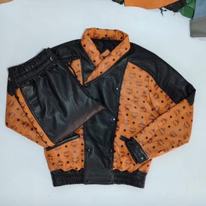 Puede incluir: Conjunto de cuero de dos piezas con chaqueta y pantalón a juego. La chaqueta tiene un cuerpo negro con mangas y cuello estampados en naranja. El pantalón es negro con una raya naranja. El conjunto es de cuero.