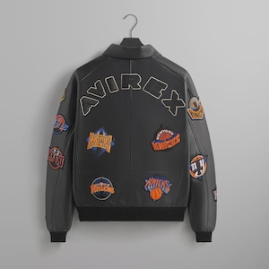 May include: A black leather jacket featuring the word AVIREX arched across the upper back in large lettering. The jacket is decorated with multiple embroidered New York Knicks basketball team patches in orange, blue, and white on the back and sleeves.