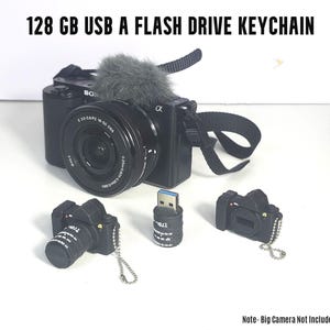 Pode incluir: Chaveiros USB em forma de câmera preta, com uma câmera maior e um microfone ao fundo. O texto diz "128 GB USB A FLASH DRIVE KEYCHAIN".