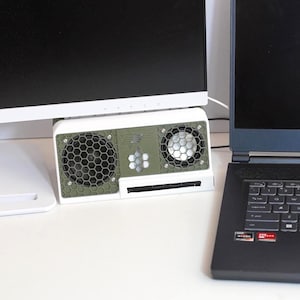 May include: A rectangular desktop speaker featuring a white outer frame and an olive green front panel with two circular speakers protected by a black hexagonal honeycomb grille. The centre of the panel displays a small honeycomb graphic design.