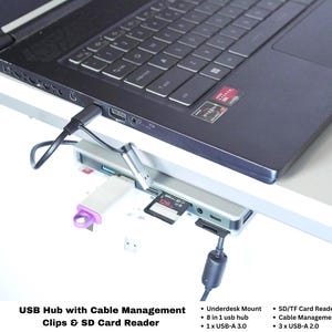 Puede incluir: Un concentrador USB 8 en 1 plateado montado debajo de un escritorio, conectado a una computadora portátil negra. El concentrador cuenta con un puerto USB-A 3.0, tres puertos USB-A 2.0 y un lector de tarjetas SD/TF. Una unidad flash blanca y una tarjeta SD están insertadas.