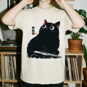 Könnte beinhalten: Ein cremefarbenes T-Shirt mit einer grafischen Illustration einer großen schwarzen Katze mit großen Augen, die an einer Computertastatur sitzt. Das Design enthält die japanischen Schriftzeichen 電腦貓 in Schwarz und ein kleines orangefarbenes Katzensymbol.