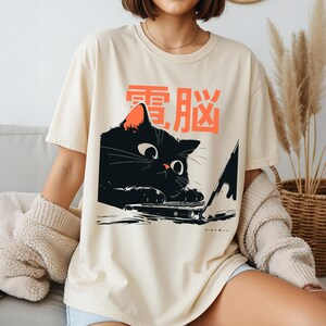 Op de afbeelding: Een crèmekleurig t-shirt met een afbeelding van een zwarte kat die naar een laptop kijkt. Boven de kat staat de oranje Japanse tekst "電脳" gedrukt. De kat heeft grote ogen en haar pootjes rusten op het toetsenbord van de open laptop.