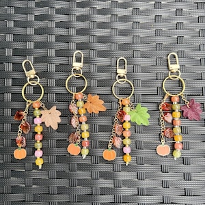 May include: Four gold-toned metal keychains featuring autumn-themed charms including orange pumpkins, various colored leaves, and strands of multi-colored beads in shades of pink, yellow, orange, and green, all attached to a circular ring and lobster clasp.