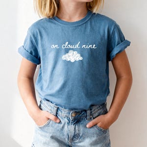 May include: A blue short-sleeved t-shirt featuring the white text "on cloud nine" written in a cursive script above a simple white line-drawn cloud graphic. The shirt has a crew neckline and is paired with light blue denim shorts with rolled cuffs.