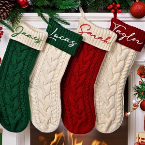 Op de afbeelding: Vier kabelgebreide kerstsokken in groen, crème en rood hangen boven een open haard. Elke sok heeft een crèmekleurige manchet met een naam geborduurd in een contrasterende kleur: James, Lucas, Sarah en Taylor. Feestelijke vakantiedecoratie omringt de sokken.