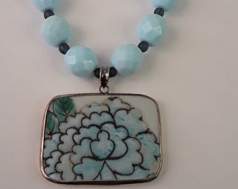 May include: A light blue beaded necklace with a silver clasp and a rectangular pendant. The pendant features a floral design in shades of blue and green, set against a white background. The necklace is a delicate piece of jewelry.